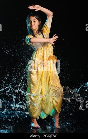 Ragazza adolescente dai capelli scuri, bella e allegra, snella, di 12 anni in un abito di seta giallo a piedi nudi in un aquazone nero. Emozioni e danza dentro Foto Stock