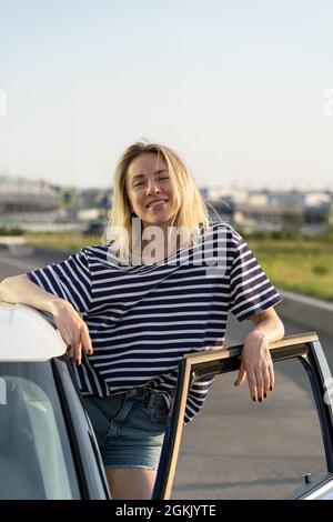 Chiosco femminile rilassato alla porta aperta dell'auto al parcheggio. Donna felice proprietario di veicolo pronto per il viaggio su strada Foto Stock