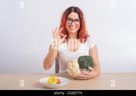 La donna caucasica preferisce cibo sano. Ragazza rossa sceglie tra broccoli e ciambelle su sfondo bianco. Foto Stock