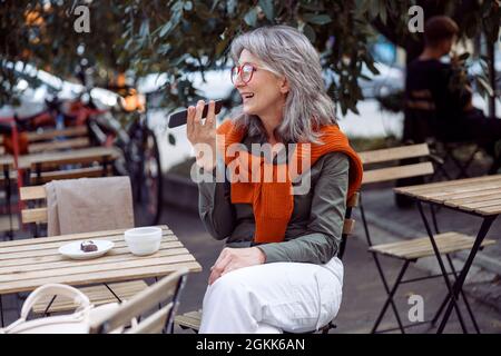 Signora con capelli d'argento con occhiali utilizza la modalità altoparlante sul telefono sulla terrazza esterna caffè Foto Stock