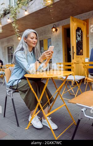 Attracive capelli lunghi Senior Asian signora tiene il telefono cellulare seduto al tavolo giallo Foto Stock