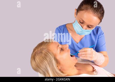 Donna bionda seduta in una sedia in studio dentista e donna stomatologo in una maschera medica e guanti protettivi esamina la cavità orale. Foto Stock