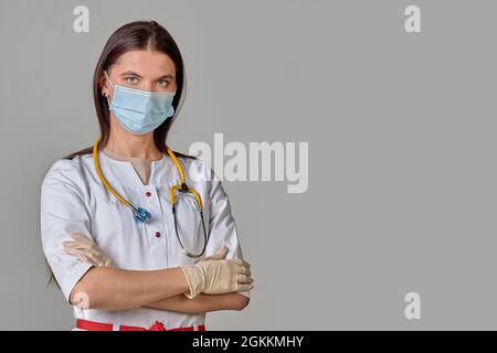 Una giovane dottore in un cappotto bianco con uno stetoscopio e una maschera medica su sfondo grigio isolato Foto Stock