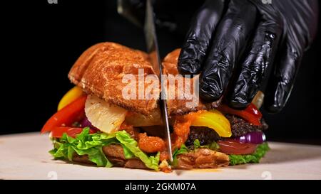 L'hamburger artigianale tagliato sta cucinando su sfondo nero in guanti da cibo neri. Consiste: Salsa di salsa rossa, lattuga, cipolla rossa, sottaceti, formaggio, peperoncino verde Foto Stock