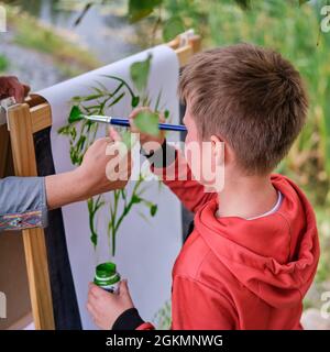 La madre insegna a ragazzo figlio di dipingere su tela. Donna insegnante artista dipinge con un bambino su carta natura e alberi presso il fiume Foto Stock