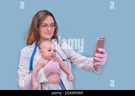 Medico pediatra che tiene il telefono e il neonato, sfondo blu studio. Infermiera felice in uniforme con un bambino e un telefono cellulare Foto Stock