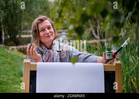 La donna felice si attinge al cavalletto con un pennello e vernici. Donna artista disegna la natura e gli alberi sulla carta vicino l'acqua sulla riva del fiume Foto Stock