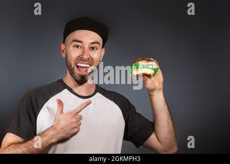 Il concetto di fast food ed ecologia. Un uomo in un cappello e una barba punta felicemente ad un hamburger nella sua altra mano. Sfondo nero. Spazio di copia. Foto Stock