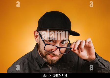 Persone ed emozioni. Un uomo con una barba e occhiali, con un cappuccio e una camicia neri, si fissa attraverso i suoi occhiali. Primo piano. Sfondo arancione. Foto Stock