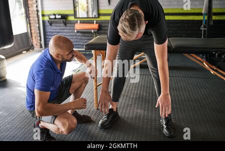 Addestramento personale che aiuta l'uomo maturo nel fare una stirata verso il basso. Assistenza per il recupero e la mobilità. Esercitarsi in palestra domestica Foto Stock