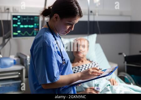 Assistente medico che controlla il trattamento dell'uomo anziano che legge le note sulla clipboard. Ammalato anziano che si trova a letto discutendo con l'infermiere circa l'esperienza di diagnosi con la sacca per infusione attaccata. Foto Stock