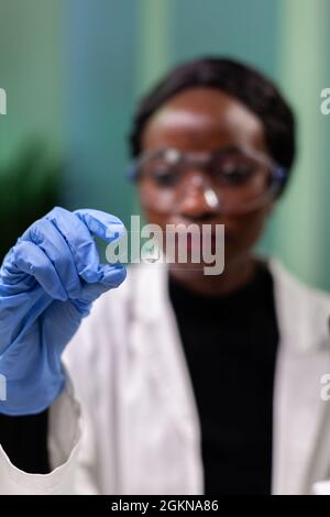 Primo piano della donna microbiologa afroamericana che guarda il campione di foglie verdi che lavora all'esperimento medico nel laboratorio ospedaliero di microbiologia. Medico botanico che analizza piante geneticamente modificate Foto Stock