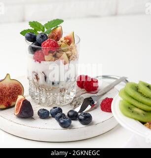 bicchiere trasparente con granola versata con yogurt, su lamponi maturi, mirtilli e fichi su tavola bianca. Colazione sana Foto Stock