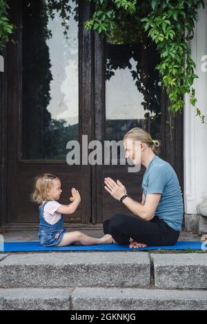 Padre e figlia che fanno yoga insieme all'aperto nel parco vicino al vecchio edificio. Una famiglia felice trascorre del tempo insieme. Concetto di stile di vita sano. Foto Stock