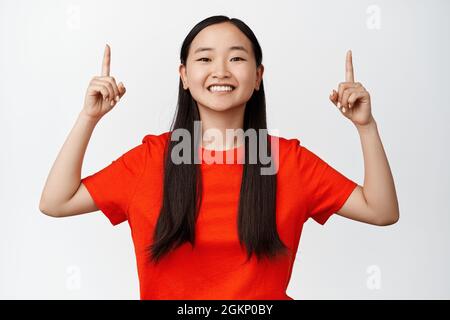 Ritratto di sorridente ragazza sorridente sorridente senso, teen asiatico puntando le dita su e guardando felice, annunciando grande vendita, in piedi su sfondo bianco Foto Stock