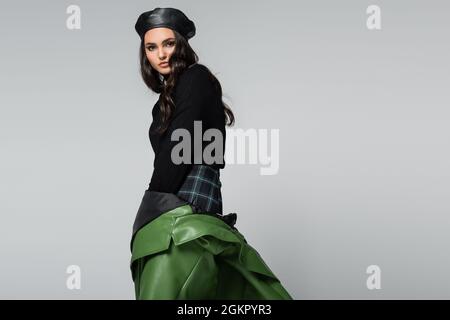 donna brunetta in gonna a plaid, berretto nero e giacca in pelle verde in posa isolata sul grigio Foto Stock