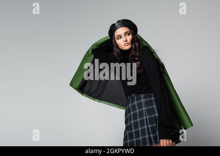 giovane donna brunetta in gonna a plaid, berretto nero e giacca in posa isolata sul grigio Foto Stock