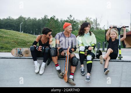 Ragazzo con scooter e tre pattinatori adulti che si rilassano sul ponte dello skatepark, godendosi. Tutti guardando il ragazzo. Vista frontale. Natura in backg Foto Stock