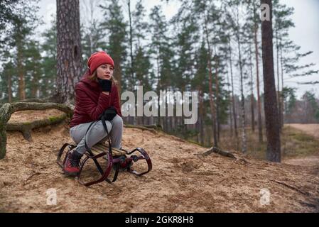 Una ragazza in una giacca rossa si siede con una slitta su una montagna nella foresta e aspetta la neve Foto Stock