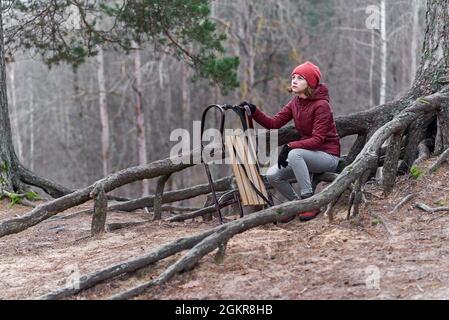 Una ragazza in una giacca rossa si siede con una slitta su una montagna nella foresta e aspetta la neve Foto Stock