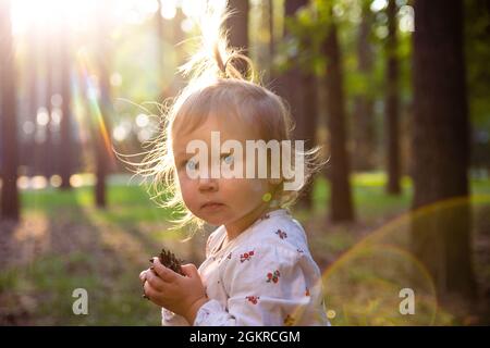 Una simpatica e adorabile bambina caucasica sta camminando in una pineta tra gli alberi in una giornata di sole al tramonto. Il sole splende attraverso capelli delicati, glar Foto Stock