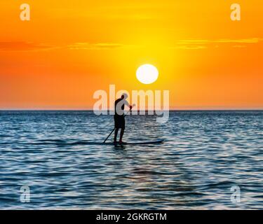 Un uomo si affaccia sul mare su una tavola da paddleboard in piedi, costeggiato dal sole che tramonta, Westward ho!, Devon, Regno Unito Foto Stock