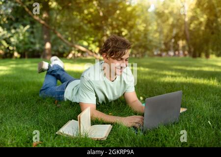 Allievo allegro che fa i compiti con il laptop e il libro, riposante nel campus universitario all'aperto, disteso sul prato Foto Stock