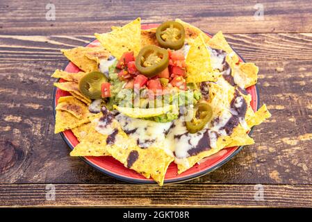 Nachos di mais messicano con guacamole, fagioli, chipotle, jalapenos, pomodoro rosso tritato su piatto rosso e tavola di legno Foto Stock