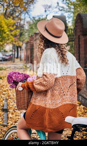 Vista posteriore della giovane donna elegante in bicicletta con bouquet di fiori in cesto. Donna bicyclist che indossa cardigan e cappello in maglia mentre si ha bicicletta all'aperto il giorno d'autunno. Foto Stock