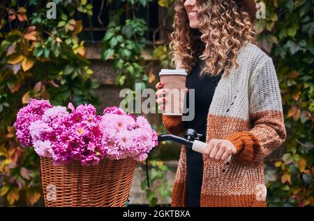 Vista laterale della donna curly in posa con la bicicletta d'epoca e una tazza di caffè vicino alla parete verde della pianta. Donna che indossa cardigan marrone è in viaggio in bicicletta con un grande cestino di camomiles rosa. Foto Stock