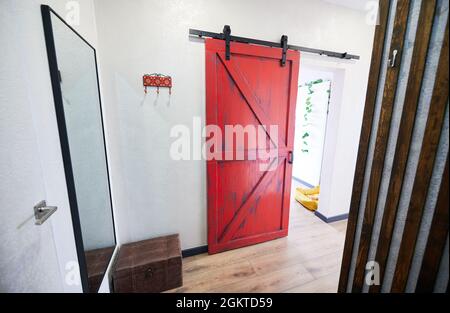 Vista della porta rossa nel corridoio con grande specchio sul muro e ottomano. Interno del corridoio dell'appartamento in stile loft con pavimento in legno. Concetto di decorazione della casa. Foto Stock