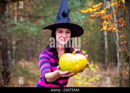 La strega di Halloween con una zucca magica in una foresta scura. Bella giovane donna in streghe cappello e costume con zucca intagliata. Halloween Foto Stock