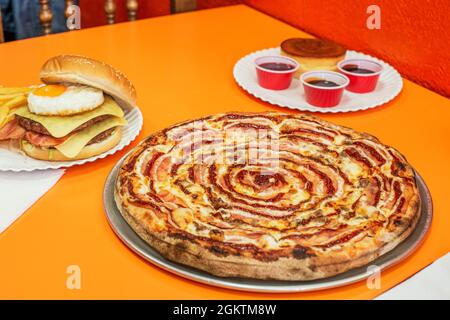 Pizza a conduzione familiare appena preparata con salsa barbecue e pancetta, accanto a un doppio hamburger al formaggio con uova fritte e patate Foto Stock