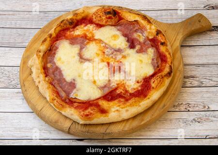 Tipica pizza italiana di pasta di grano sottile con prosciutto e mozzarella gratinati su un asse di legno Foto Stock