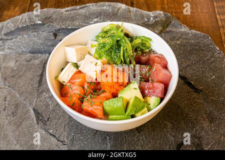 Ciotola di poke con uova di pesce, salmone, tonno rosso, avocado verde, tofu bianco e alghe guacamole con semi di sesamo su sfondo di pietra grigia Foto Stock