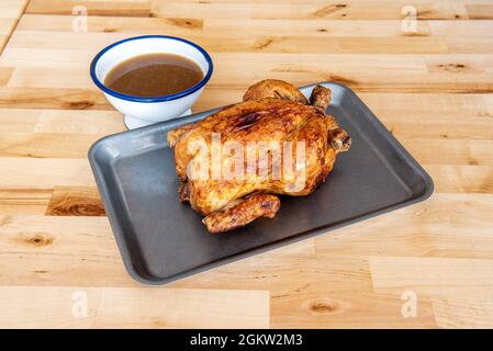 Pollo arrosto intero con la sua pelle croccante su vassoio di metallo e succo di cottura in un recipiente separato su tavolo di legno Foto Stock