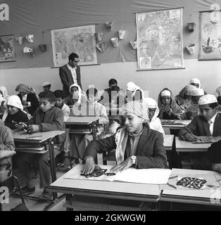 anni sessanta, ragazzi storici, giovani, alcuni con i headdresses sopra, seduti in una classe ad una scuola primaria, con insegnante maschile, tagliare e fare forme e modelli da carta. Foto Stock