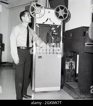 Anni '60, storico, un tecnico televisivo in uno studio televisivo, accanto ad una macchina di riproduzione o proiettore RCA TP66, Jeddah, Arabia Saudita. Il TP-66 di RCA fu introdotto nel 1964 e divenne ampiamente utilizzato nelle stazioni televisive di tutto il mondo per la riproduzione di film sonori silenziosi e magnetici da 16 mm, sia in diretta in aria che in videocassetta per l'editing o la trasmissione differita. Usato anche per la riproduzione di una stazione commerciale, sono stati un workhorse di industria progettato per eseguire 24/7, riproducendo il contenuto del film che ha costituito molto programma televisivo nei primi 30 anni della televisione. Foto Stock