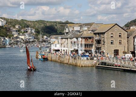 Un'affollata banchina all'East Looe con un'attraente barca a vela rossa che percorre il fiume Looe in Cornovaglia. Foto Stock