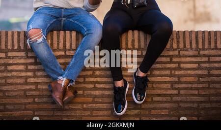 Coppia seduta su muro romano mattoni indefinito. Uomo in jeans blu e stivali marroni, donna in leggings neri e sneakers, parete arancione come panca Foto Stock