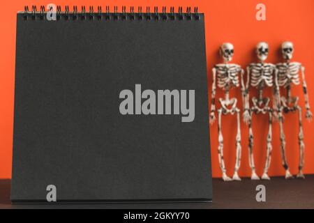 Notebook nero vuoto su sfondo arancione con scheletri sfocati. Design minimalista per le feste di Halloween. Mock up. Copia spase. Foto Stock