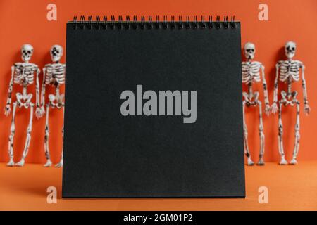 Notebook nero vuoto su sfondo arancione con scheletri sfocati. Halloween moderno o concetto di giorno dei morti. Un design minimalista per le vacanze. Foto Stock
