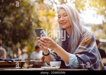 Allegra donna asiatica senior scrive sms su smartphone seduto al tavolo Foto Stock