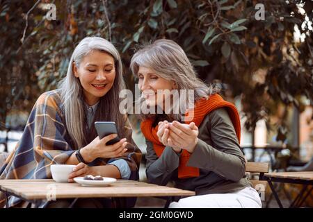 Sorridente donna asiatica mostra foto al telefono ad un amico seduto in Street cafe Foto Stock