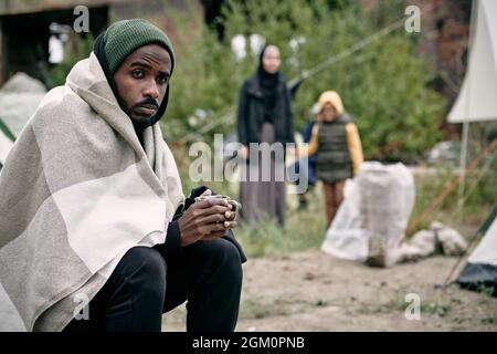 Puzzled giovane uomo nero avvolto in coperta seduta nel campo migrante e bere il tè in tempo freddo Foto Stock