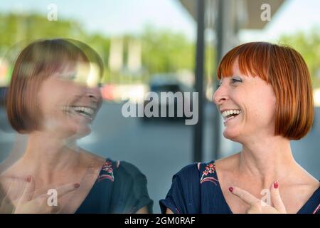 Gioiosa giocosa attraente donna rossa matura ridendo e indicando il suo riflesso in una grande finestra di vetro di lastra di un edificio commerciale in un ur Foto Stock