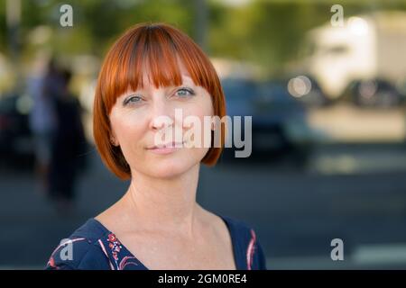 Donna di mezza età pensierosa e attraente rossa che guarda la macchina fotografica con un'espressione intensa e un sorriso tranquillo in una strada urbana verdeggiante in primo piano Foto Stock