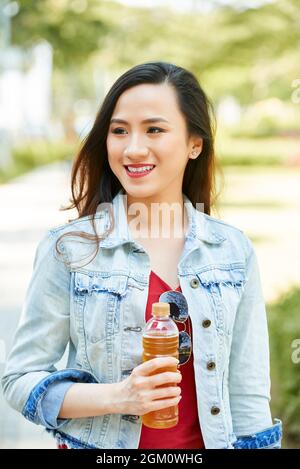 Ritratto di bella donna giovane positiva con bottiglia di plastica di bevanda rinfrescante camminando nel parco Foto Stock