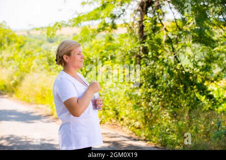 Foto del profilo di una donna sportiva anziana in piedi e riposata dopo il jogging mattutino nel parco, cerca di aprire una bottiglia d'acqua che sta tenendo dentro Foto Stock