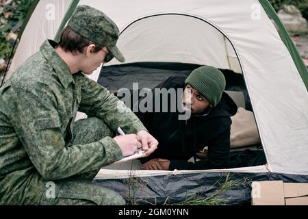 Soldato in uniforme dell'esercito accovacciato alla tenda dell'uomo nero mentre esegue il conteggio dei migranti Foto Stock